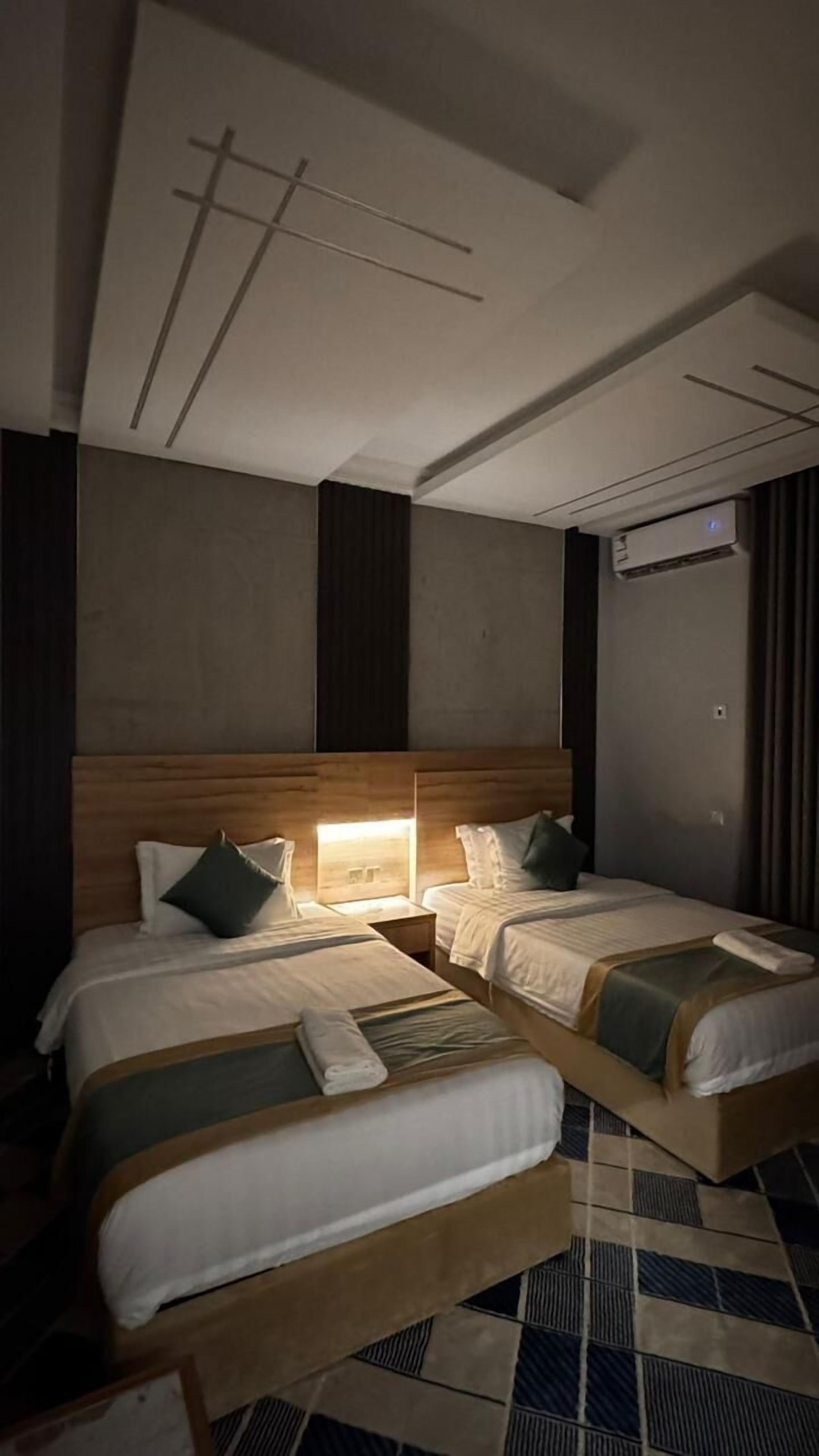 Ett hotellrum med två sängar, en sänggavel i trä, ett mönstrat golv och ett tak med infälld belysning.