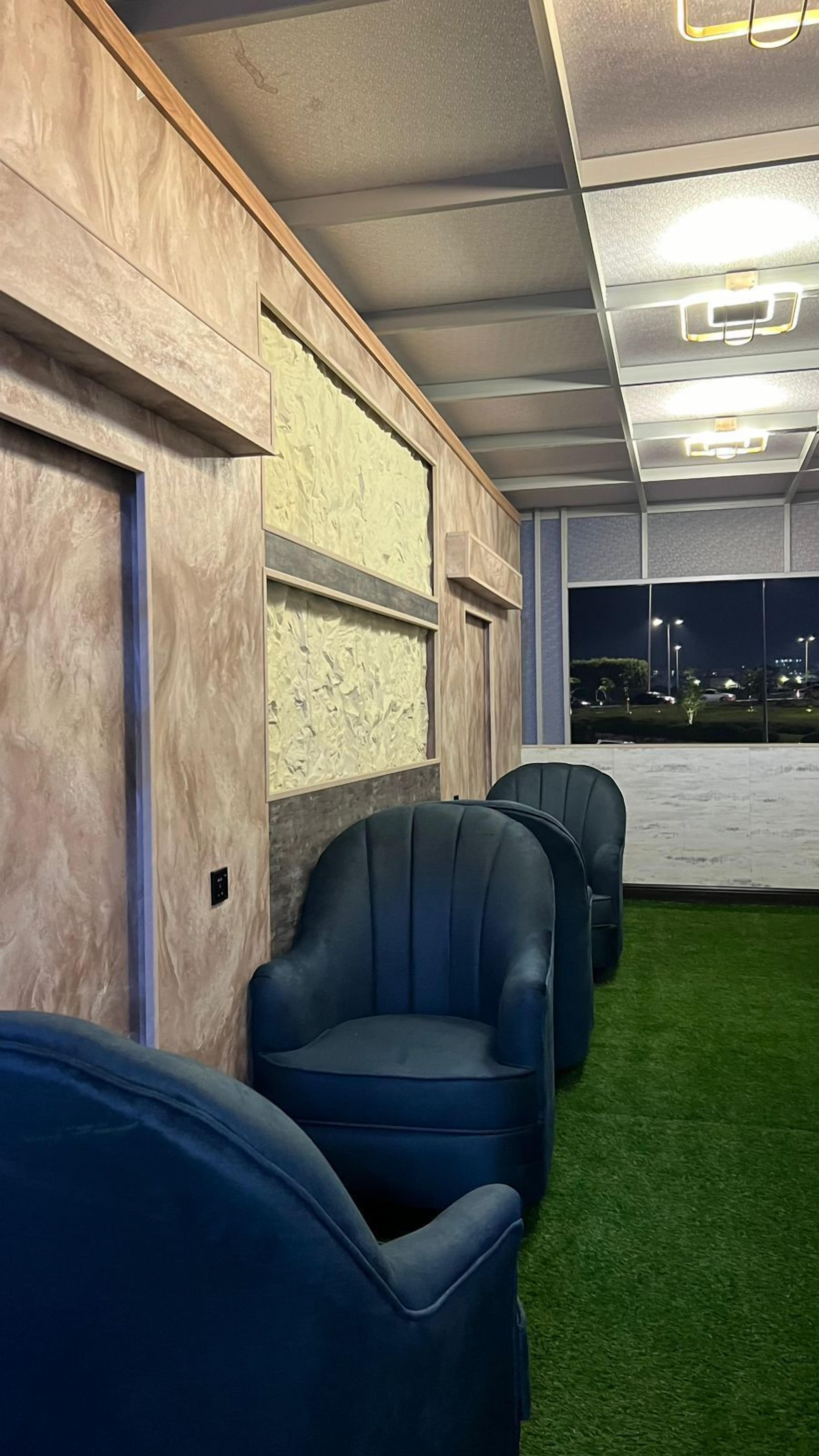 Fila de sillas azules sobre suelo de césped artificial verde, pared de madera con panel texturizado e iluminación empotrada en el techo.