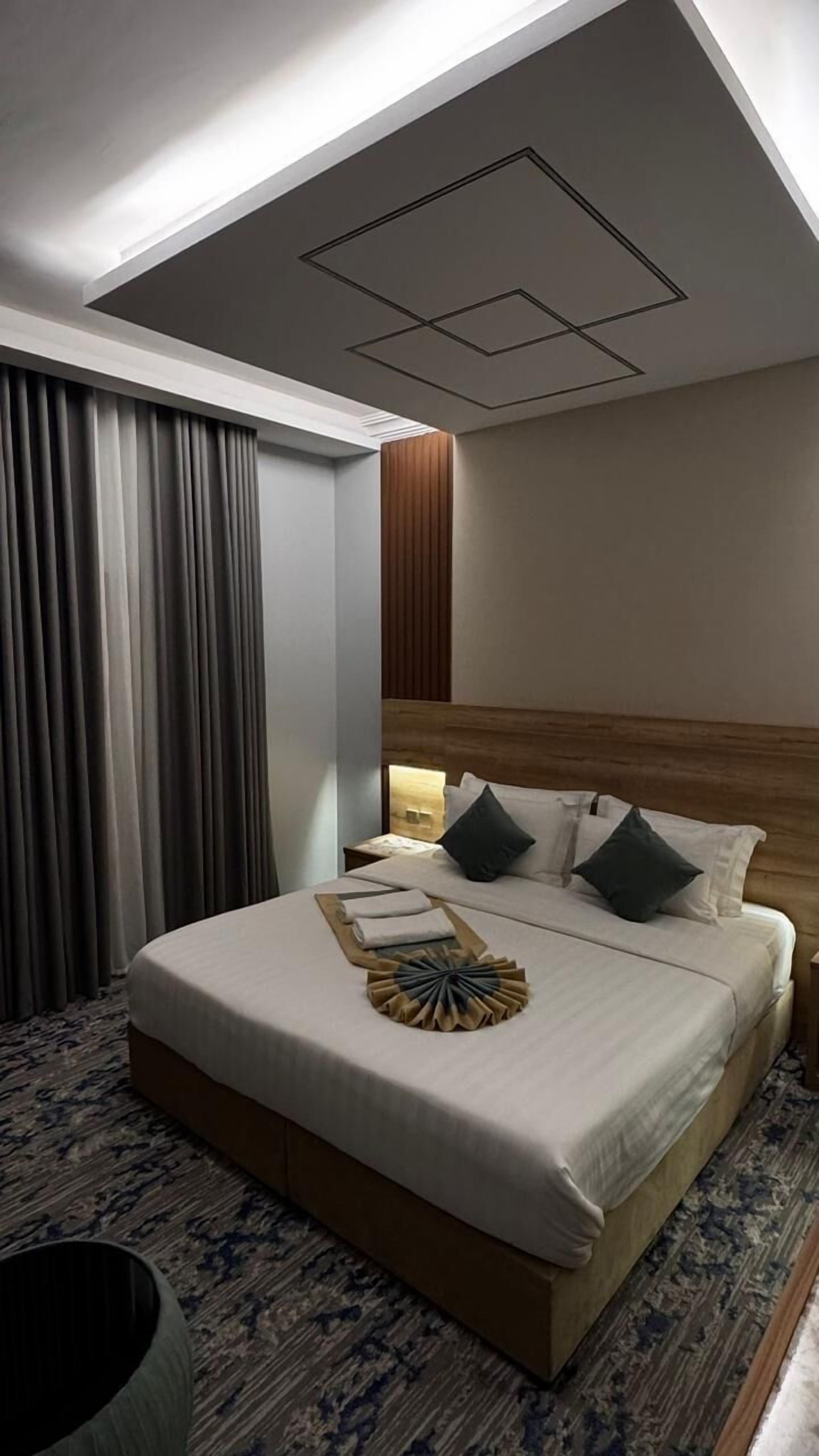 A hotel room with a large bed, a wooden headboard, a nightstand with a lamp, and a ceiling with recessed lighting.