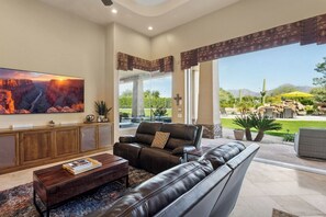 A spacious living room with a large window, leather sofas, a coffee table, and a mounted TV. - Lavish Estate w/THREE Sport Courts & Home Theater (Scottsdale)