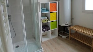 Casa de banho com chuveiro em cabine de vidro, armário branco com gavetas coloridas, um banco de madeira e janela que proporciona luz natural.