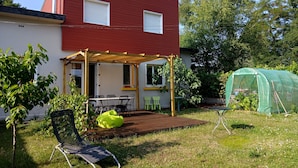 Uma casa de dois andares com fachada vermelha, um alpendre coberto com mesa e cadeiras, e uma área de jardim verde com um banco e uma pequena estufa.