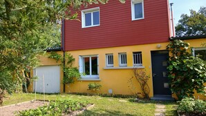 Uma casa de dois andares com telhado vermelho e paredes amarelas, uma porta da frente e um pequeno jardim com plantas.