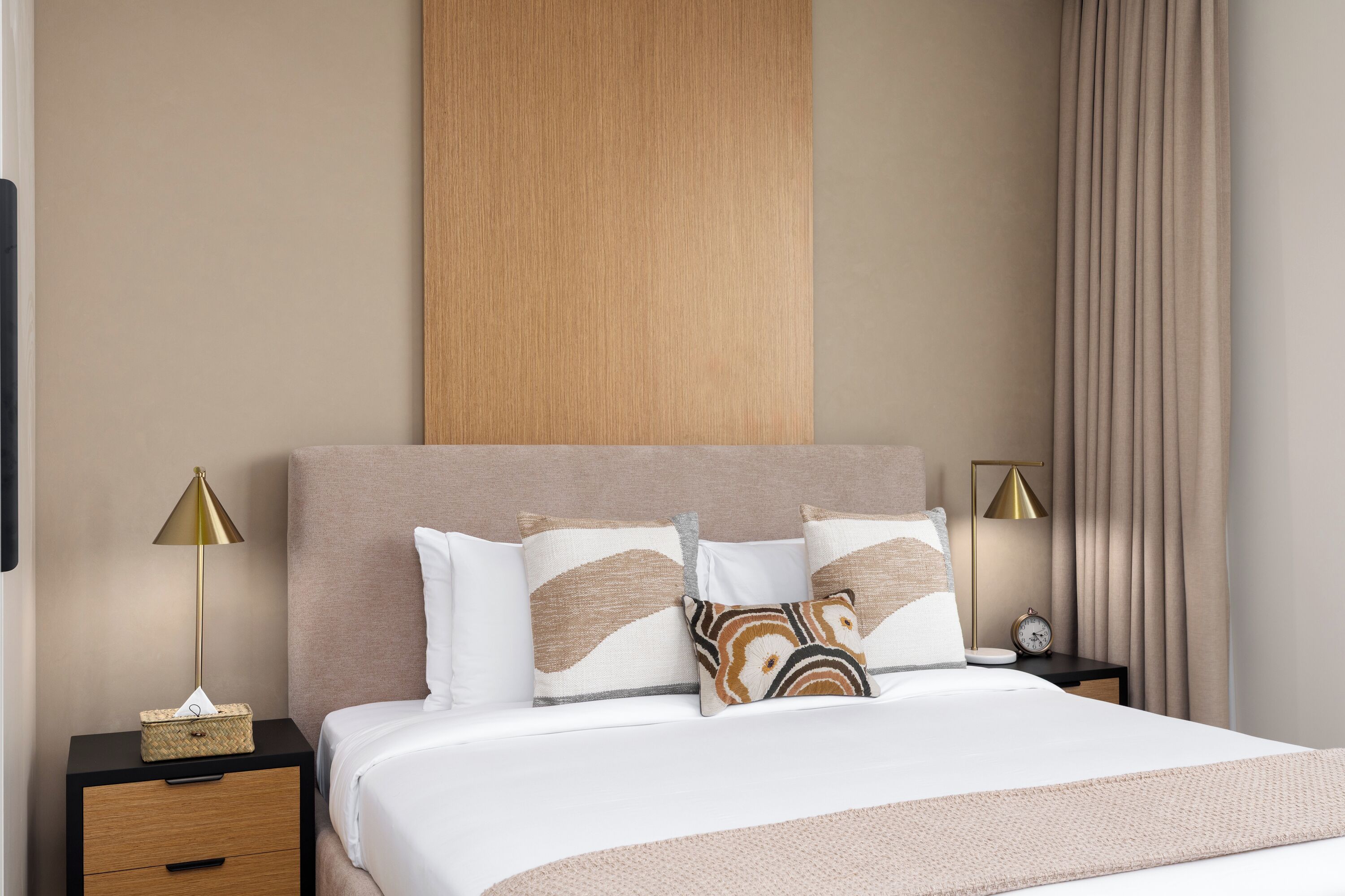A neatly made bed with a white comforter and decorative pillows, a wooden headboard, a bedside table with a lamp, and a nightstand with a clock.