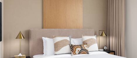 A neatly made bed with a white comforter and decorative pillows, a wooden headboard, a bedside table with a lamp, and a nightstand with a clock.