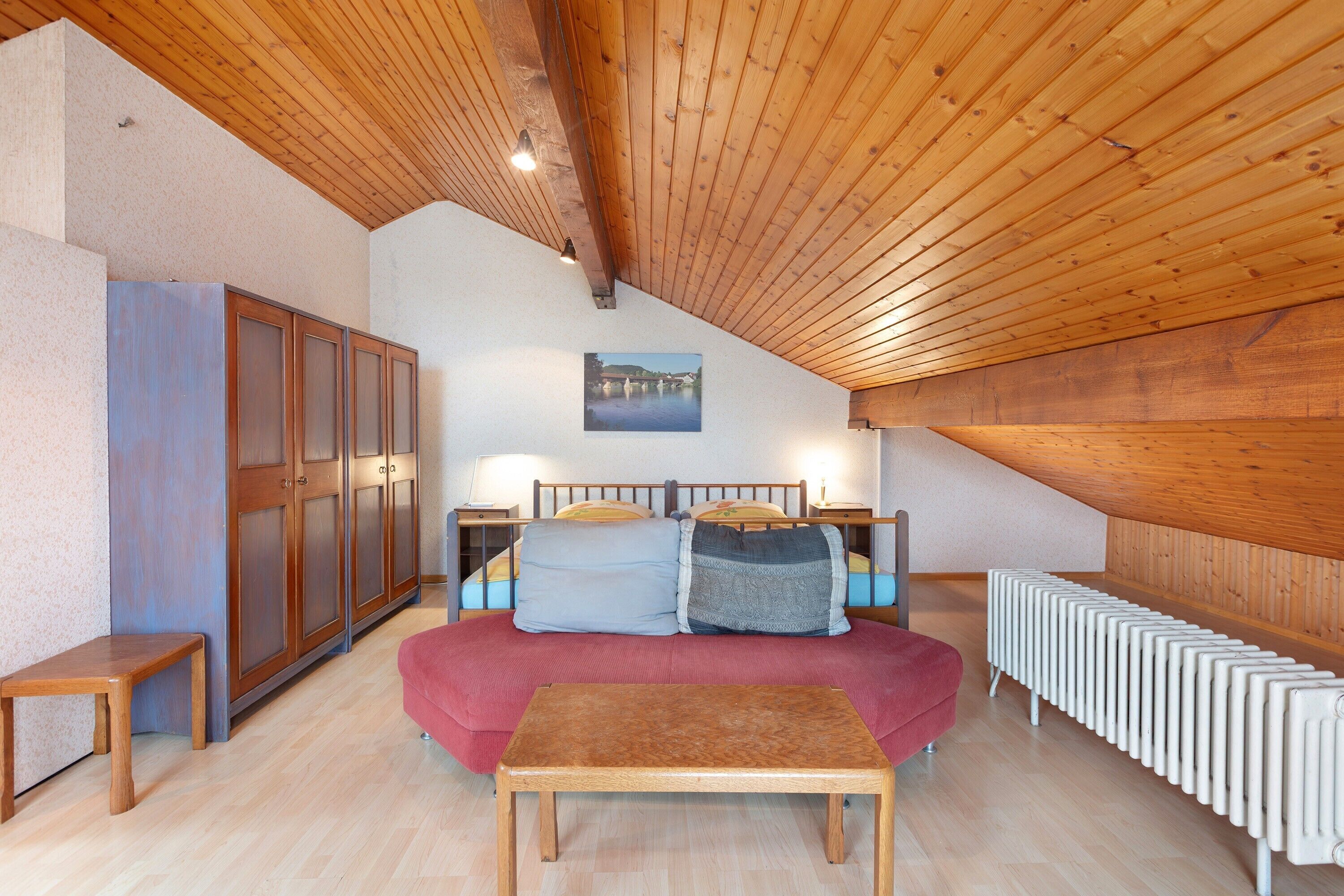 Ein Schlafzimmer mit Holzbalkendecke, einem Bett mit Kissen, einem Nachttisch, einer Bank und einer Heizung.
