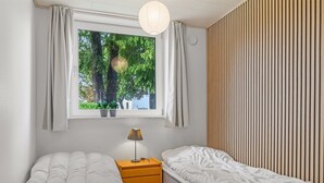 Un dormitorio con dos camas individuales, una mesita de noche con lámpara, una ventana con cortinas y vistas a árboles.