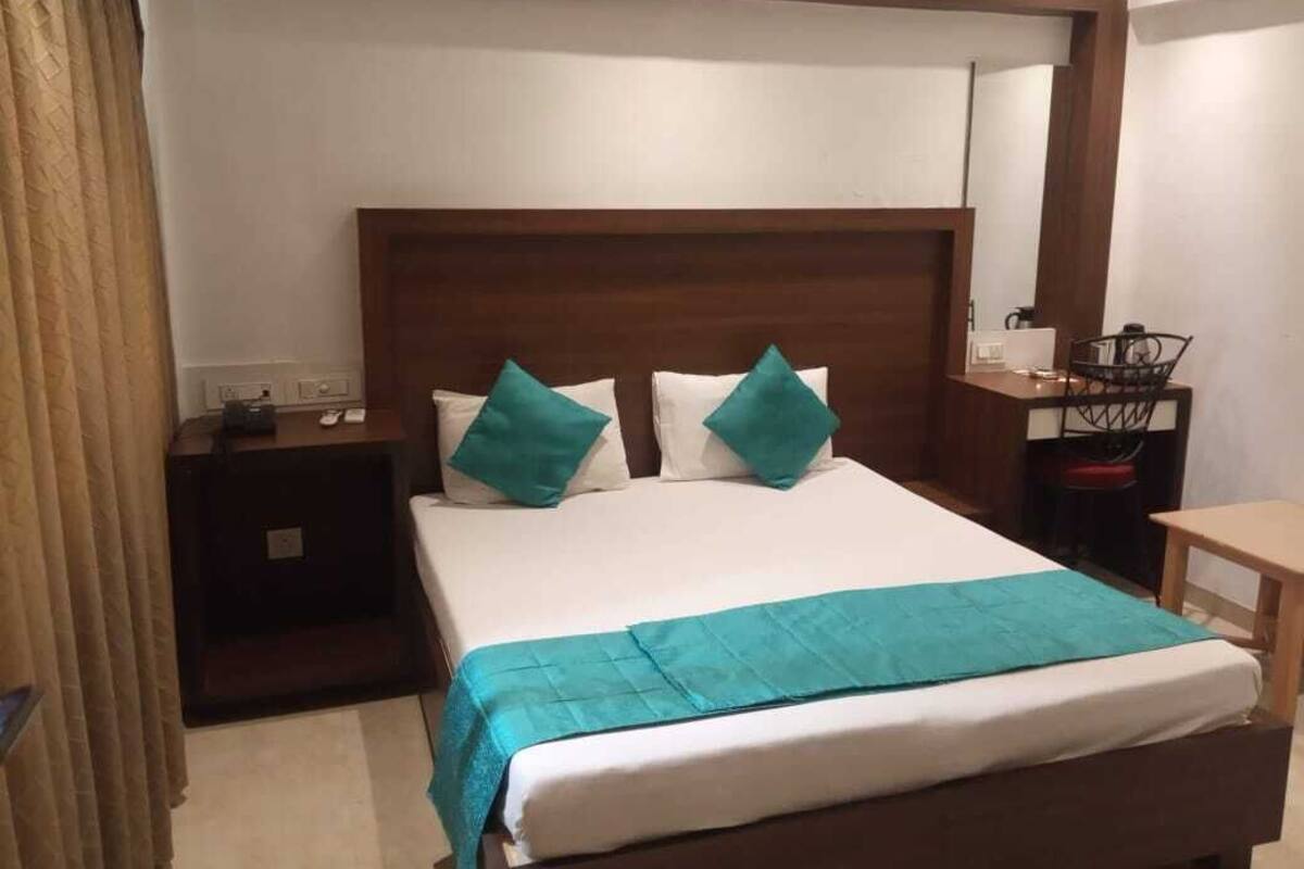 A hotel room with a wooden headboard, a bed with white bedding and teal pillows, a bedside table with a lamp, and a small desk with a chair.