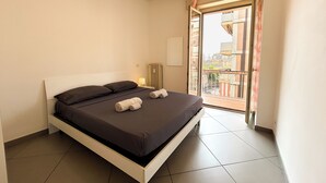 Un lit bien fait, avec une literie sombre et deux serviettes enroulées, une lampe de chevet et une vue sur un balcon avec une balustrade et des bâtiments en arrière-plan.