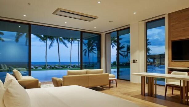 A bedroom with a large bed, a sofa, a dining table, and a TV. It has large windows with a view of palm trees and the ocean. - Saba Estate Luxury Villa Bali (Bali)