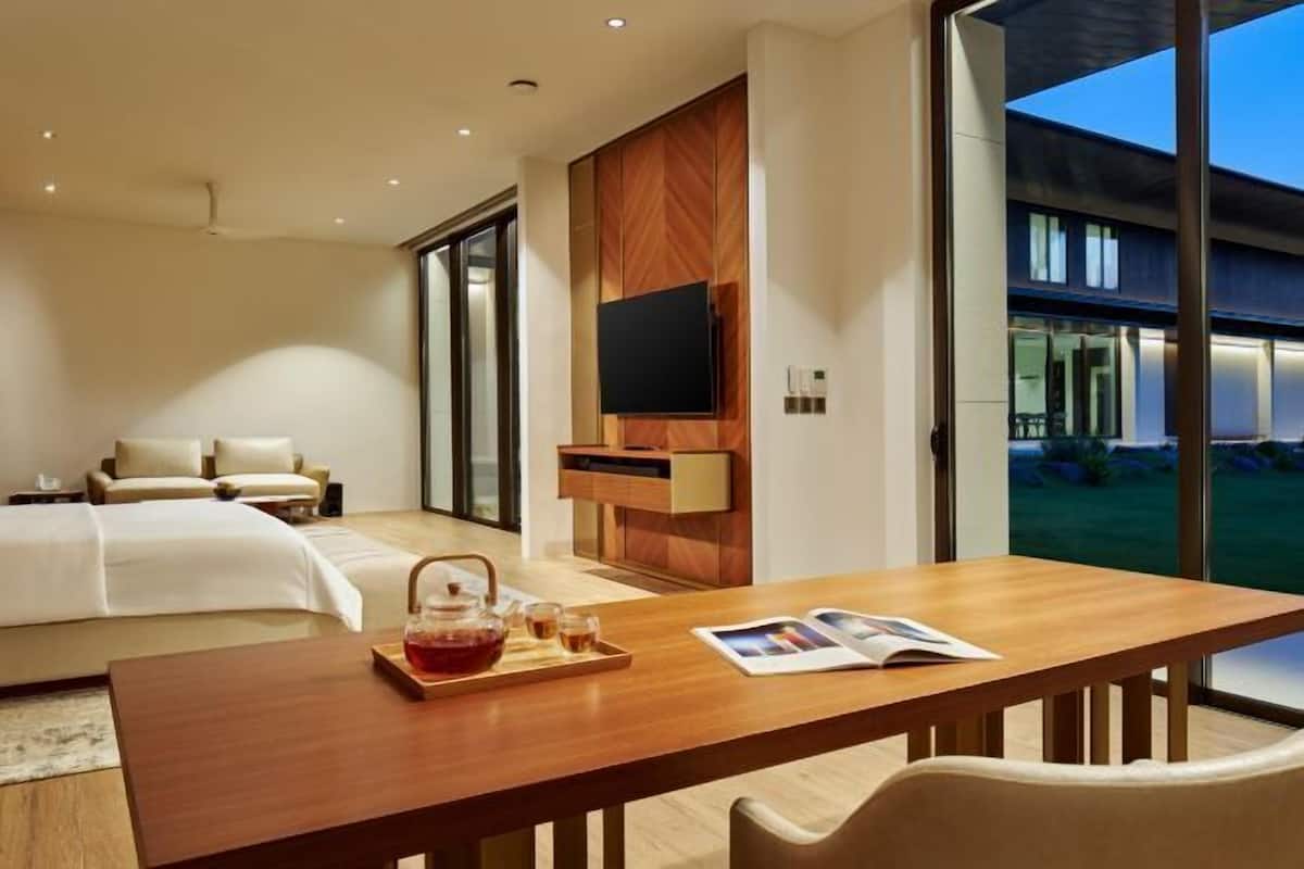 A modern hotel room with a large bed, a wooden table with a tray of tea, a flat-screen TV mounted on a wooden panel, and a view of the outdoors through large windows.