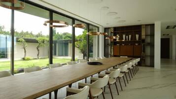 Una larga mesa de comedor con sillas, ventanas amplias y vistas a un jardín.
