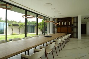 Una larga mesa de comedor con sillas, ventanas amplias y vistas a un jardín.