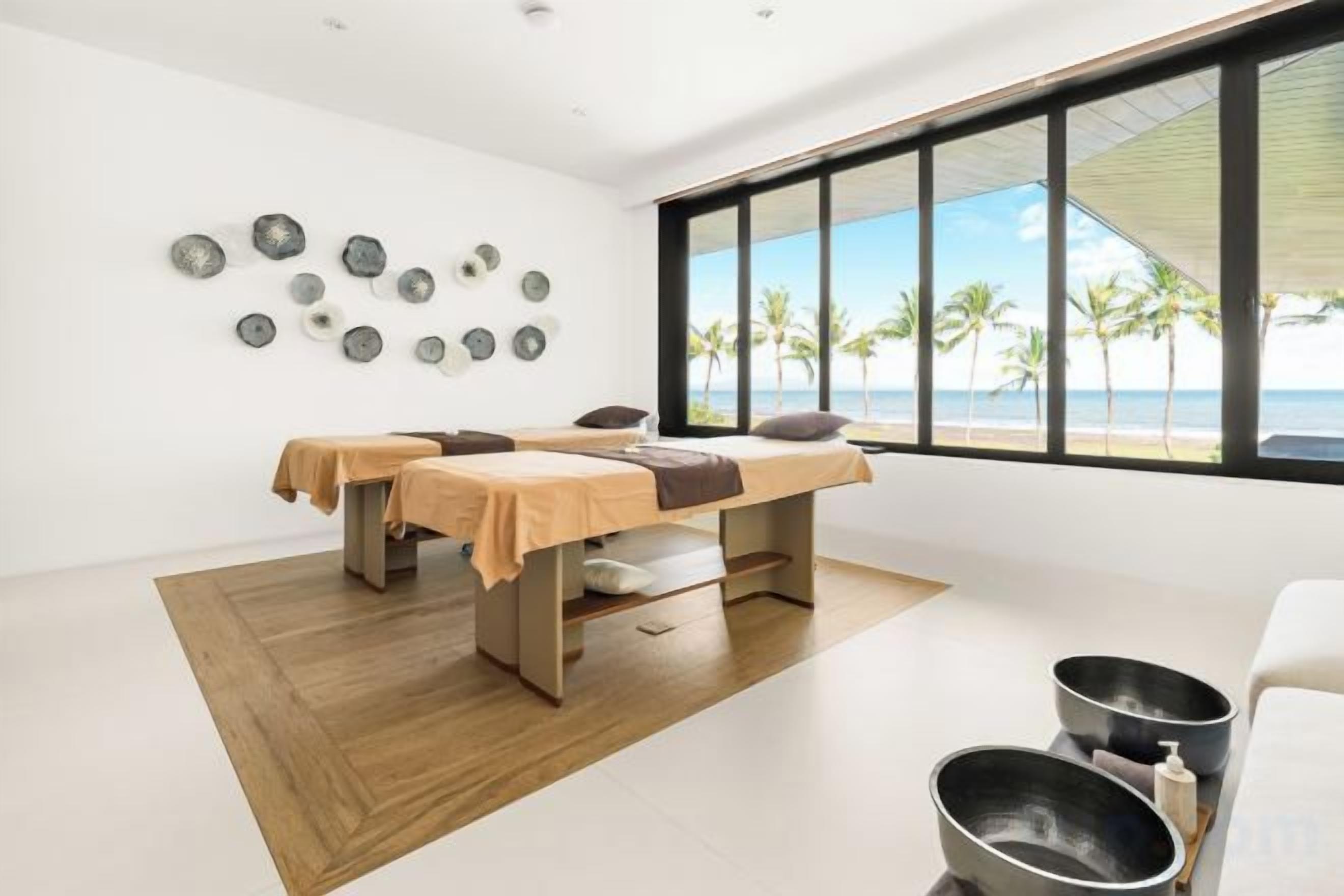 Un cuarto de spa con dos camillas de masaje, un ventanal con vista a la playa y suelo de madera.