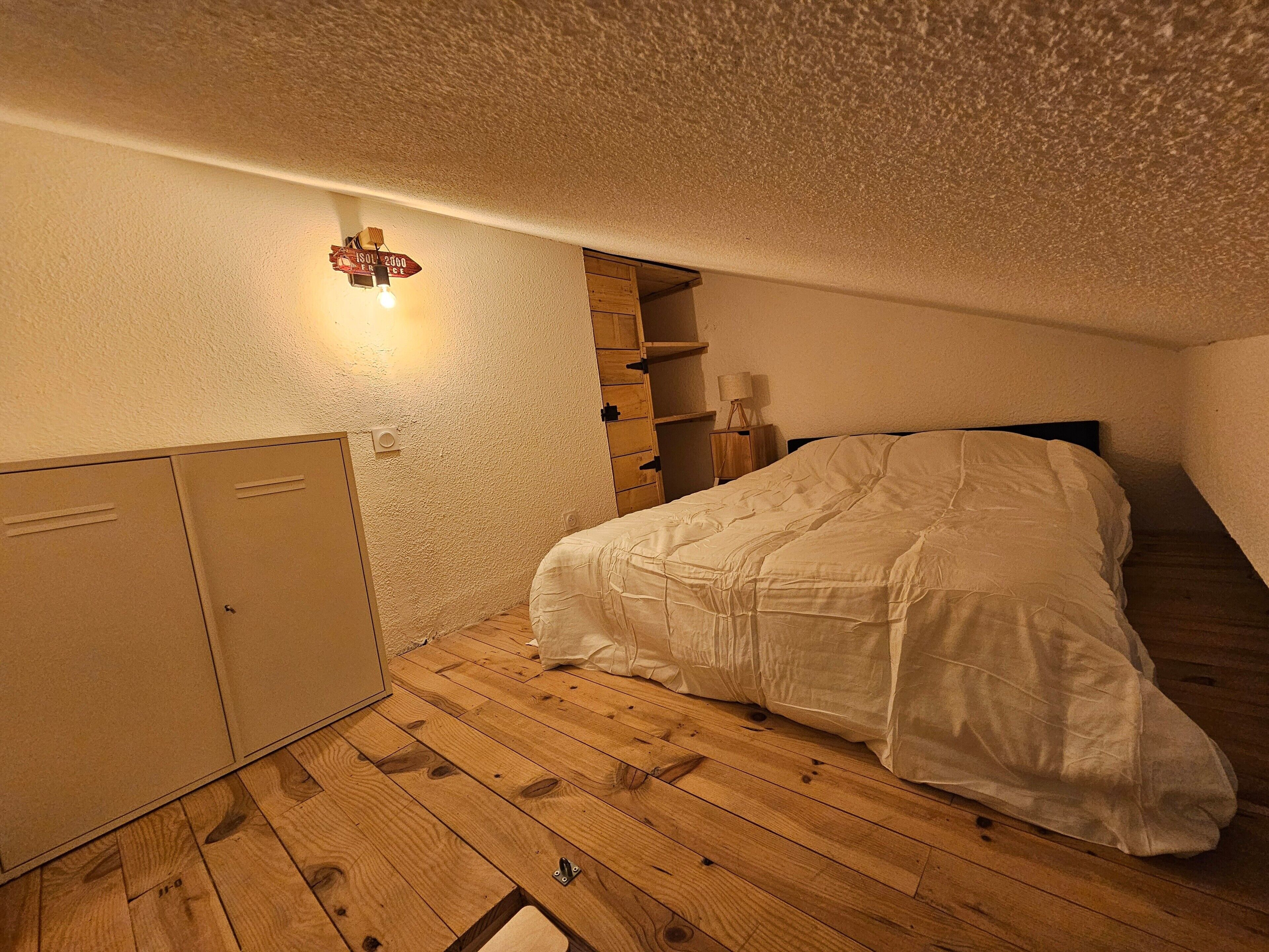 Ett sovrum med en säng, trägolv, en garderob, en hyllsektion och en väggmonterad lampa.