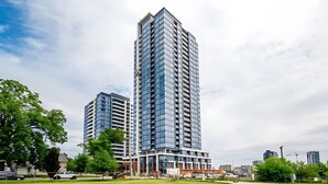 A modern high-rise building with a glass facade, surrounded by a well-maintained lawn and other buildings in the background. - Stylish 1BR Condo w Balcony Skyline Views (Kitchener)