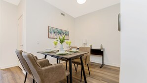 A dining area with a table set for four, surrounded by chairs, a painting on the wall, and a small side table with a lamp and decorative items. - Stylish 1BR Condo w Balcony Skyline Views (Kitchener)