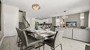 A modern kitchen with white cabinets, a dining area with a table set for four, and a grey sofa in the living area. - Upstay Championsgate 9BR Villa With Pool Cinema (Davenport)