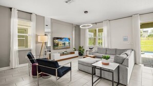 A modern living room with a grey sofa, a black chair, a coffee table, and a TV mounted on the wall. - Upstay Championsgate 9BR Villa With Pool Cinema (Davenport)