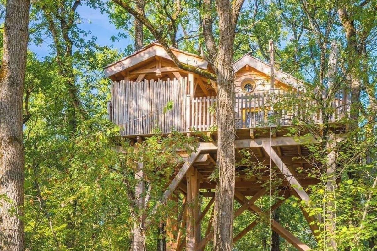 Ein Baumhaus mit Holzterrasse und rundem Fenster, umgeben von dichtem Wald.