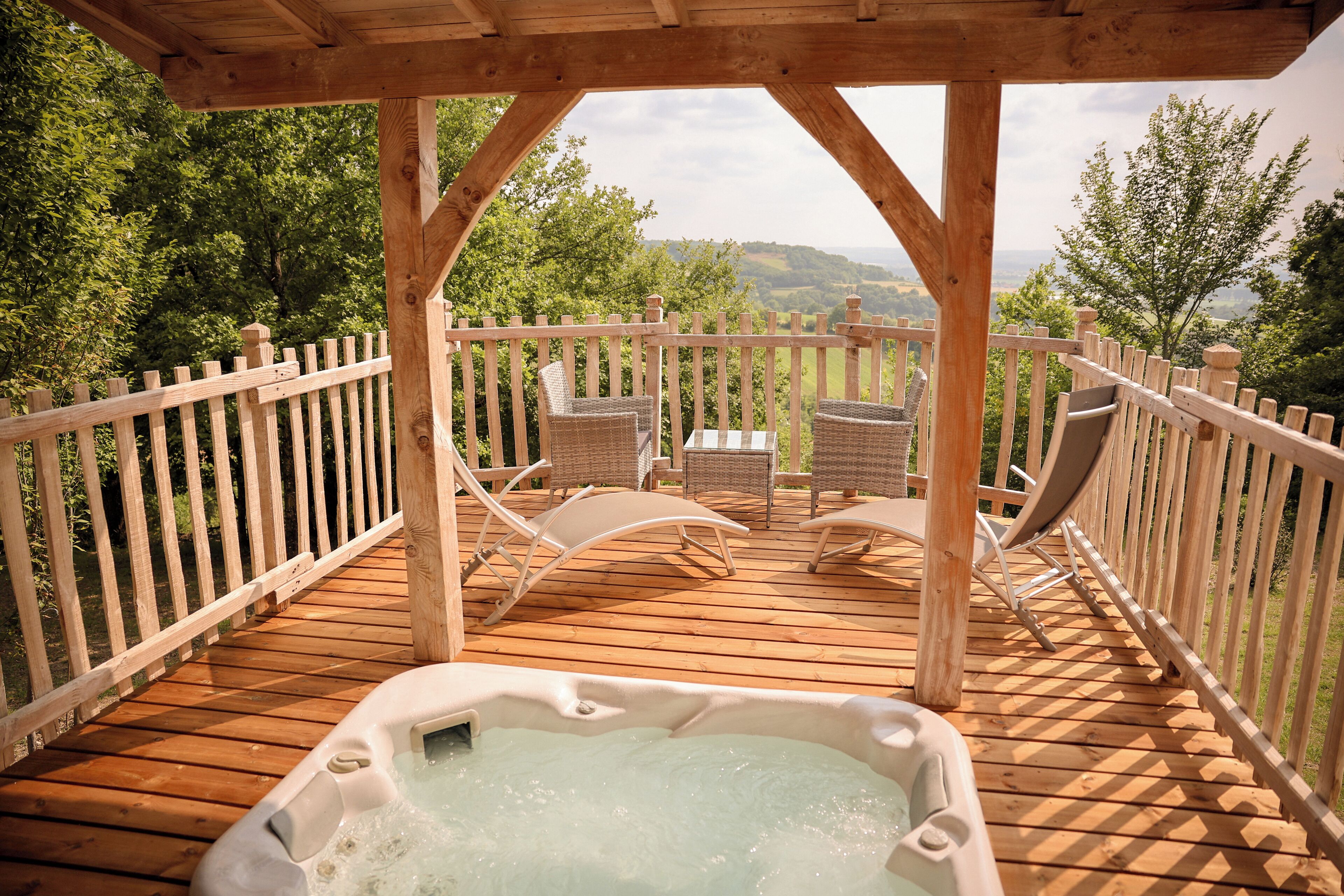 A wooden deck with a hot tub, wicker chairs, and a wooden railing overlooking a scenic landscape.
