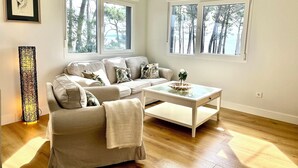 A modern living room with a beige sofa, a coffee table, and a floor lamp. There are two windows with a view of trees.