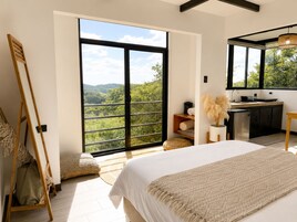 Un dormitorio con un ventanal grande, una cama, una mesita de noche, un espejo y una kitchenette.