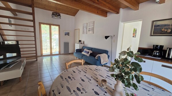 A cozy living room with a blue sofa, a dining table with a fish pattern tablecloth, a coffee machine, and a large window with a view of greenery.