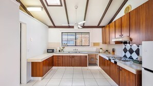 Une cuisine avec des armoires en bois, un plan de travail blanc, une fenêtre avec un grillage métallique, un ventilateur de plafond et un réfrigérateur.