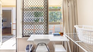A small room with a white desk, a chair, a window with a metal screen, and a laundry basket.