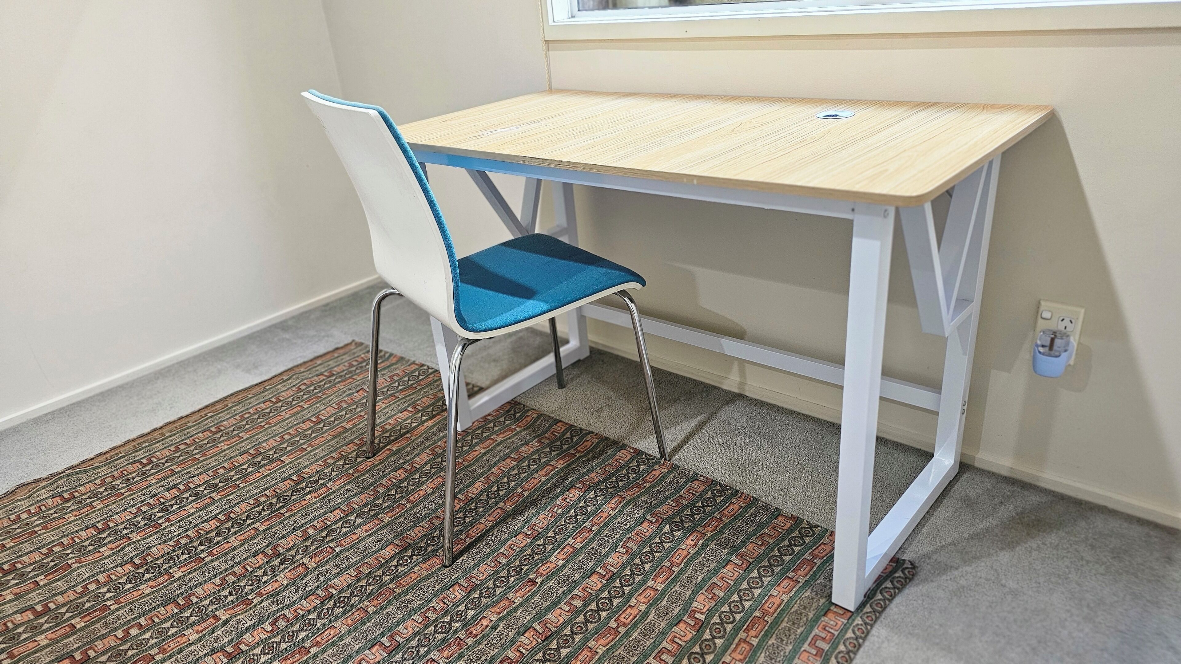 A small, white, metal-framed desk with a light wooden top, positioned next to a white chair with a blue seat cushion. The room features a patterned carpet and a plain wall with a window above the desk.