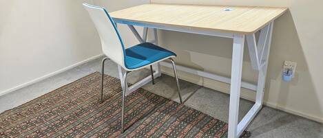 A small, white, metal-framed desk with a light wooden top, positioned next to a white chair with a blue seat cushion. The room features a patterned carpet and a plain wall with a window above the desk.