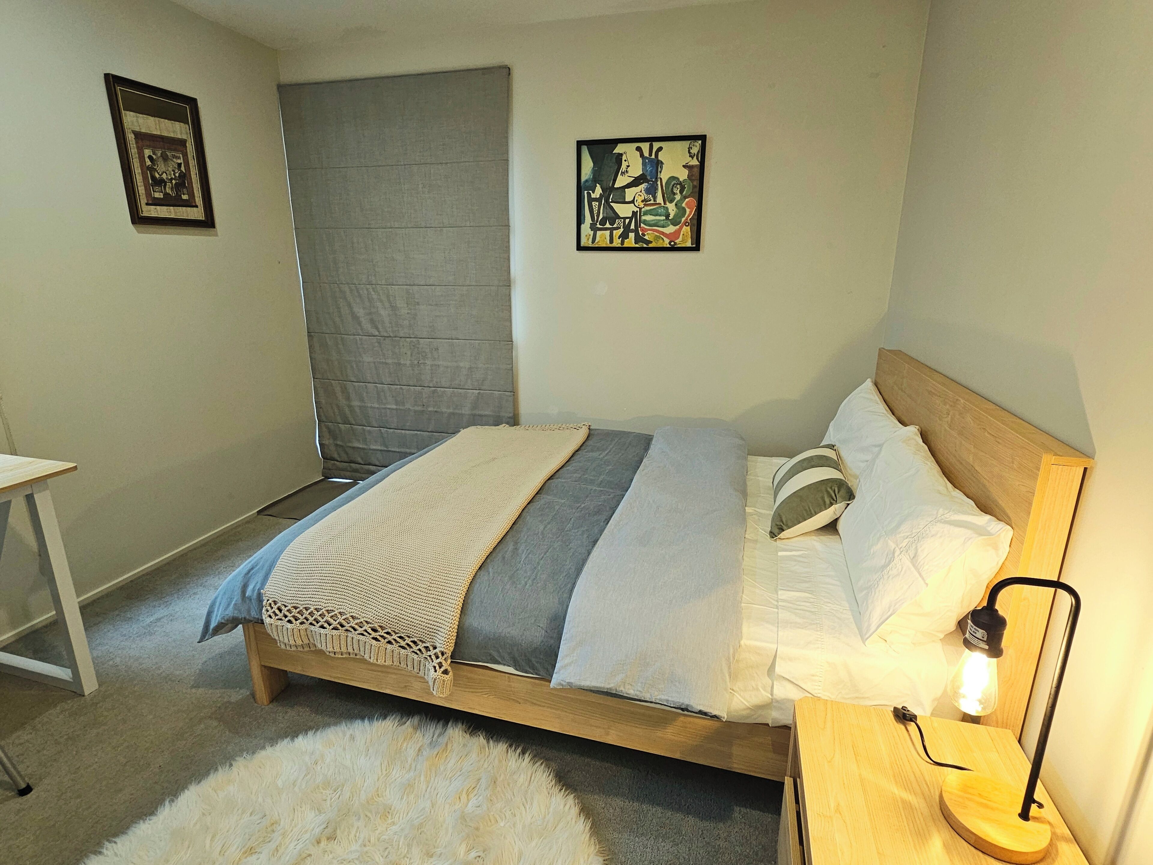 Una camera da letto con un letto, un comodino con una lampada, una piccola scrivania, un tappeto morbido e due quadri appesi al muro.