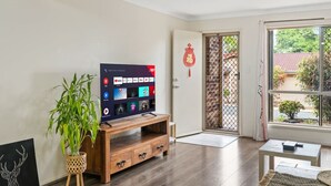 Een woonkamer met een flatscreen televisie op een houten standaard, een potplant en een deur die naar buiten leidt.
