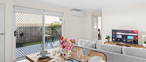 A dining area with a table set for four, a grey sofa, and a TV displaying a streaming service interface.