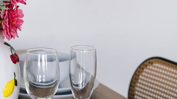 Une table à manger avec deux verres à vin transparents, un bol blanc et un vase contenant des fleurs roses.