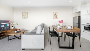 Un salon moderne avec un canapé gris, une table basse en bois et un téléviseur diffusant une chaîne YouTube. On y trouve également un coin repas avec une table en bois dressée pour quatre personnes, des chaises et un vase de fleurs. La cuisine est équipée de meubles blancs et d’un réfrigérateur.