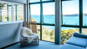 A room with a large window offering a view of a body of water and greenery. There is a white furry chair and a blue sofa.