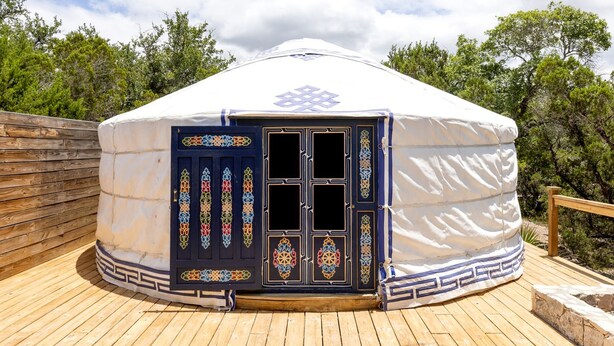Une yourte traditionnelle dotée d’une porte décorative, d’une terrasse en bois et entourée d’arbres.