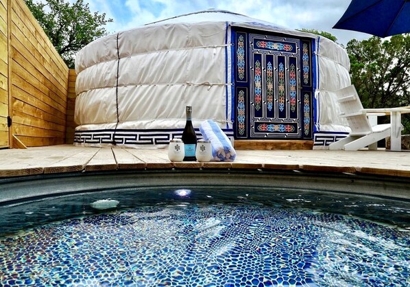 A hot tub with mosaic tiles, a white tent-like structure with a decorative door, and a bottle of champagne.