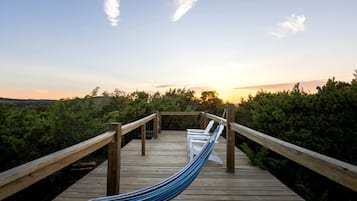 A wooden deck with a hammock, white chairs, and a wooden railing overlooking a forested area.