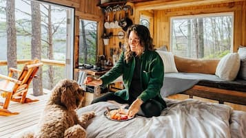 A cozy cabin interior with a large bed, a wooden ceiling, and a window overlooking a forest.