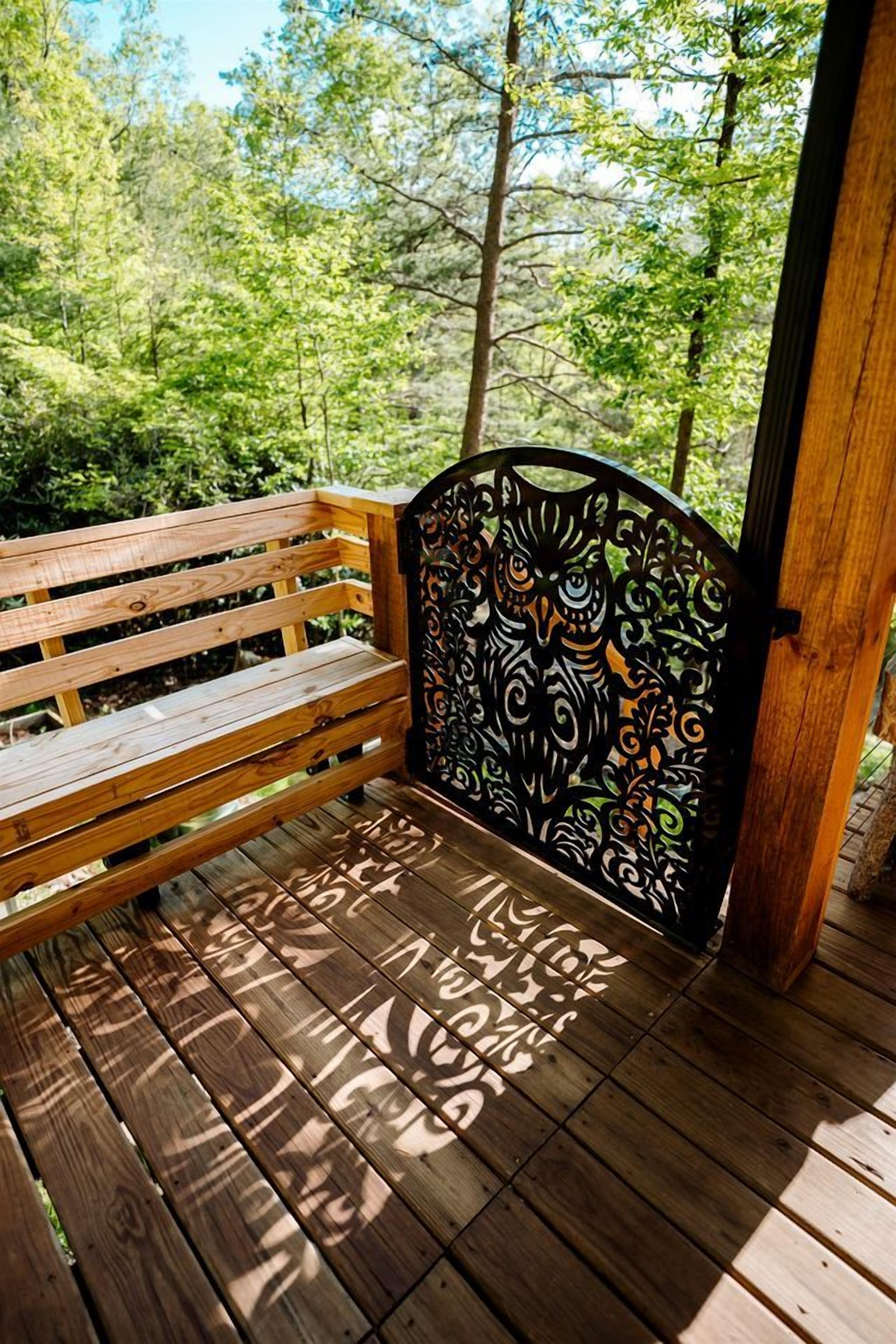 A wooden bench with a metal backrest featuring an owl design, set on a wooden deck with a forest background.