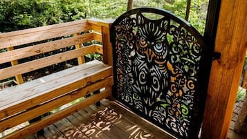 A wooden bench with a metal backrest featuring an owl design, set on a wooden deck with a forest background.