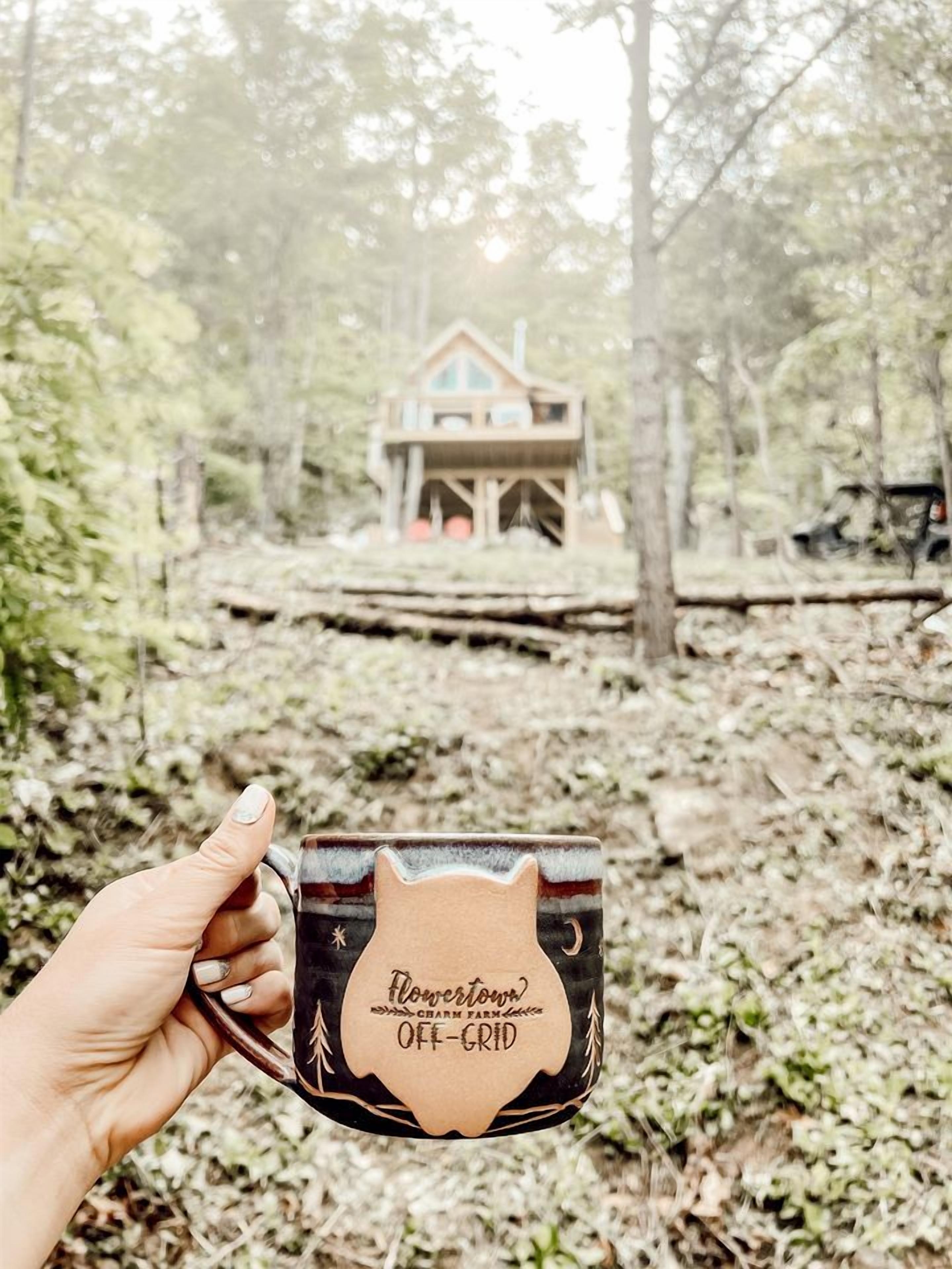 Uma mão segurando uma caneca com a inscrição 'Flower Town' e 'Charm Farm', com uma floresta e uma casa ao fundo.
