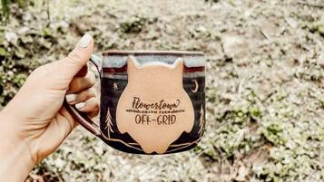 Uma mão segurando uma caneca com a inscrição 'Flower Town' e 'Charm Farm', com uma floresta e uma casa ao fundo.