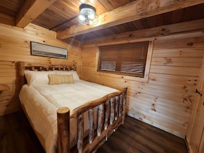 A wooden cabin bedroom with a bed, a headboard, a window with blinds, a ceiling light fixture, and a framed picture on the wall.
