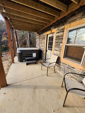 A covered patio with a hot tub, two chairs, and a stone wall.