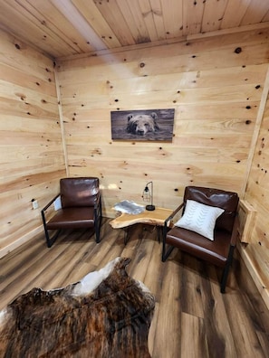 Ein Holzblockhaus-Interieur mit zwei Ledersesseln, einem Bärenbild, einem kleinen Tisch mit Lampe und einem Fellteppich auf dem Boden.