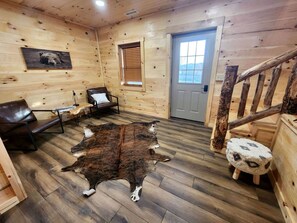 A log cabin interior with a wooden floor, a bear painting, a leather chair, a wooden stool, and a staircase.